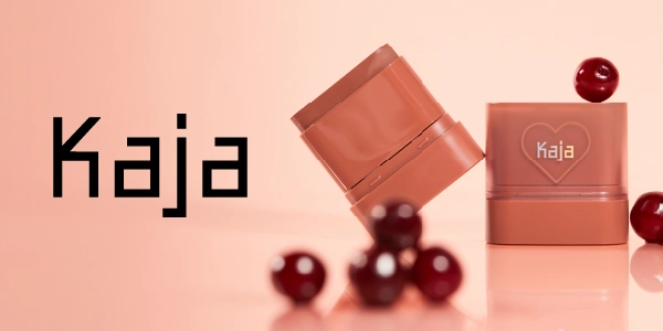 Kaja: dwa brzoskwiniowe sztyfty do makijażu, jeden z logo Kaja w sercu, na różowym tle z wiśniami.