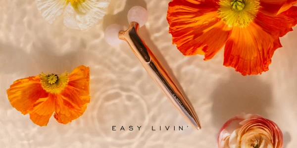 Rose gold face massager with rose quartz ends in rippled water, surrounded by orange and white poppies.