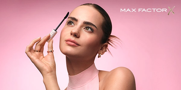 Max Factor tusz do rzęs. Kobieta unosi wzrok, aplikując mascarę na rzęsy. Zbliżenie twarzy na różowym tle.
