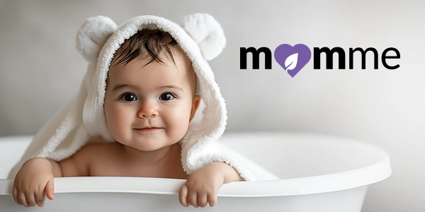 Uśmiechnięte niemowlę w białym ręczniku kąpielowym z kapturem w wannie. Logo momme z sercem i liściem.