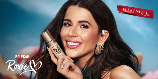 Uśmiechnięta Roxie prezentuje podkład Rimmel Multi-Tasker Better Than Filters w złotej butelce, z logo Rimmel.