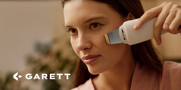 Kobieta używa białego urządzenia Garett do peelingu kawitacyjnego na policzku. Zbliżenie na twarz, metalowa łopatka.