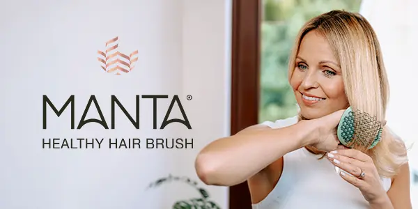 Blonde woman smiles, brushing her long hair with a flexible green Manta Healthy Hair Brush, logo on white wall.