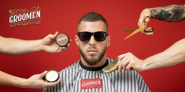 Man in barber's cape, sunglasses, surrounded by GROOMEN beard & hair products, comb, and barber scissors.