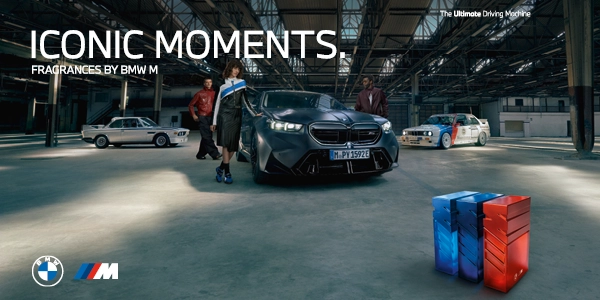 Flakony perfum BMW M, niebieski i czerwony, na podłodze. W tle klasyczne i nowoczesne auta BMW oraz ludzie w magazynie.