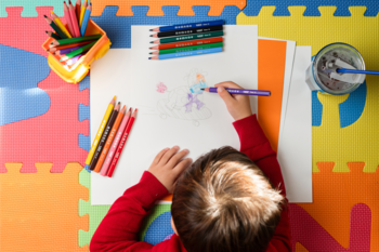 Dziecko rysuje na białej kartce, wokół kolorowe kredki i flamastry. Ujęcie z góry, nauka trzymania.