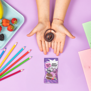 Dziecięce ręce trzymają spiralną suszoną przekąskę jabłko-malina-porzeczka, obok fioletowe opakowanie.