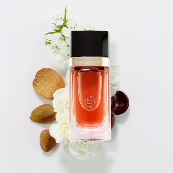 Bugatti Eleganza Ambra, flakon perfum o zaokrąglonych krawędziach, otoczony białymi kwiatami, migdałami i wiśniami na białym tle.