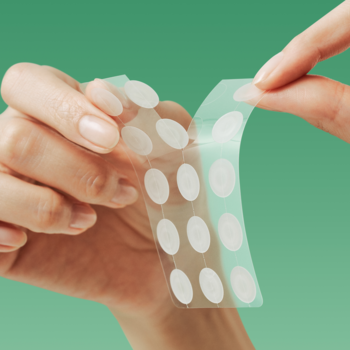 Zbliżenie dłoni odklejających arkusz półprzezroczystych, wodoodpornych plastrów na wypryski o różnej średnicy, na zielonym tle.