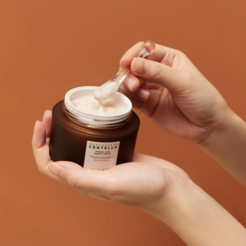 Skin1004 Probio-CICA Enrich Cream: zbliżenie na rękę nabierającą biały krem szpatułką z brązowego słoiczka.