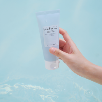 Zbliżenie ręki trzymającej jasnoniebieską tubkę Skin1004 Madagascar Centella Hyalu-Cica Sleeping Pack nad wodą.