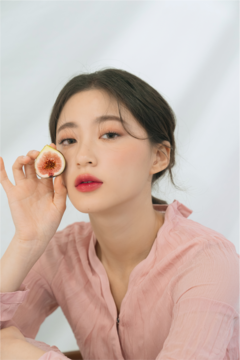 Młoda kobieta z błyszczącymi ustami, trzymająca figę przy policzku; estetyka Rom&nd Juicy Lasting Tint.