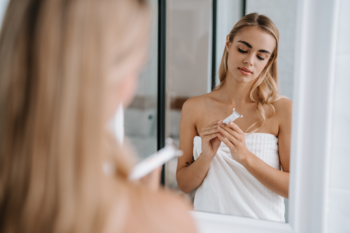 Blondynka w łazience w ręczniku patrzy w lustro z tubką, kontekst użycia golarki Beautifly My Blade Silky.