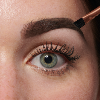 Zbliżenie na oko i brew. Pędzelek aplikuje brązową pomadę z Revolution Brow Sculpt Kit, modelując brew.
