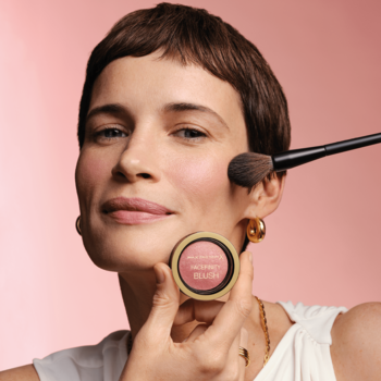 Kobieta z uśmiechem aplikuje róż Max Factor Facefinity Sunkissed Rose, trzymając złote opakowanie.