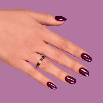 Dłoń z połyskującym śliwkowym lakierem do paznokci i pierścionkiem z kolorowymi koralikami na fioletowym tle.