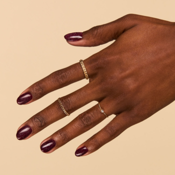 Semilac 393 Sparkling Black Cherry, brokatowy lakier hybrydowy. Dłoń z ciemnowiśniowymi paznokciami i złotymi pierścionkami na jasnym tle.