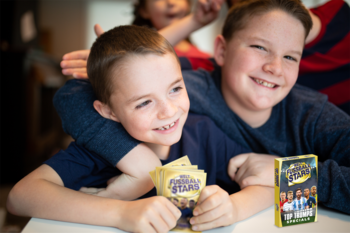 Dwóch uśmiechniętych chłopców gra w karciankę Top Trumps World Football Stars; jeden trzyma karty, obok pudełko.