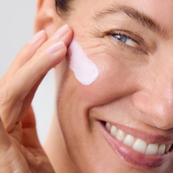 Zbliżenie na uśmiechniętą kobietę aplikującą różowy krem NIVEA HYALURON CELLULAR FILLER na policzek.