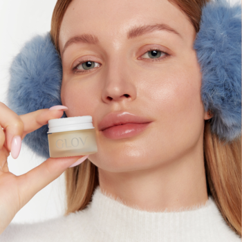 Kobieta w futrzanych nausznikach trzyma małe masełko Glov BioRetinol do ust o zapachu mango. Wygładzający wegański balsam.