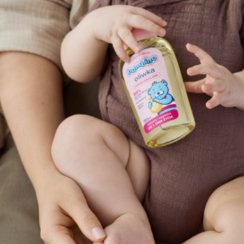 Hipoalergiczna oliwka Bambino w złotej butelce z różową etykietą, trzymana przez dziecko na rękach dorosłego.
