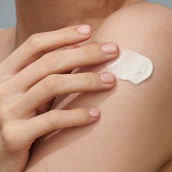 Zbliżenie na dłoń aplikującą lekki krem przeciwsłoneczny z SPF 50+ na ramię. Krem o satynowym wykończeniu.