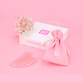 Różowy kamień Gua Sha Ilū do masażu twarzy, z woreczkiem i pudełkiem, ułożony na różowym tle.