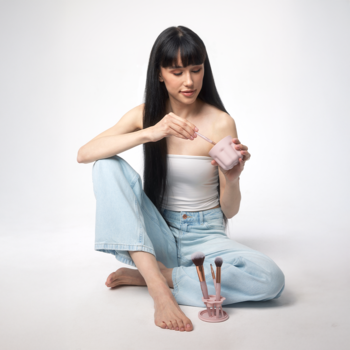 Kobieta czyści pędzel w różowej, kompaktowej myjce do pędzli. Obok stoi różowy stojak na pędzle, białe tło.