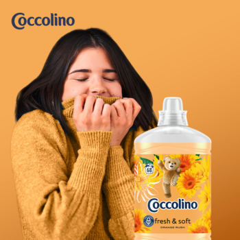 Coccolino Fresh & Soft płyn do płukania Orange Rush, kobieta w swetrze wącha tkaninę. Żółta butelka, kwiaty.