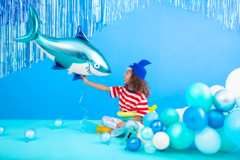 Błyszczący niebieski balon foliowy rekin PartyDeco, trzymany przez dziecko na tle imprezowych dekoracji.
