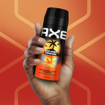Dezodorant AXE Sunset Fresh o zapachu ananasa i mango w czarnej puszce, trzymany przez dłoń na pomarańczowym tle.