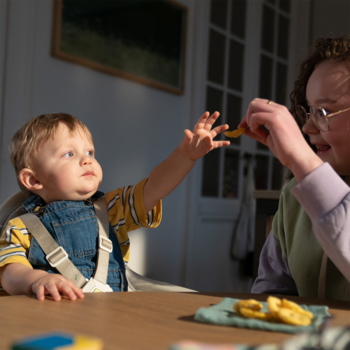 Maluch w krzesełku do karmienia sięga po owocową przekąskę podawaną przez dorosłego, scena karmienia dziecka.