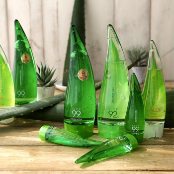 Holika Holika 99% Aloe Soothing Gel, zielone butelki w kształcie aloesu, ułożone na drewnianym tle z liśćmi.