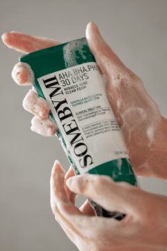 Ręce w pianie trzymają zieloną tubkę oczyszczającej pianki Some By Mi AHA BHA PHA do cery trądzikowej.