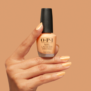 Ręka trzyma lakier OPI Beauty School Popout. Buteleczka z półprzezroczystym, brzoskwiniowym lakierem, na paznokciach ten sam kolor.