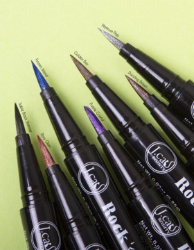 J.Cat Beauty Rock Glitz: 7 brokatowych eyelinerów w pisaku z widocznymi końcówkami, ułożonych na zielonym tle.