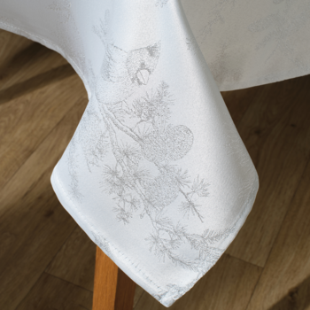 Biały obrus z delikatnym szarym wzorem sosnowych gałęzi i szyszek, zwisający z rogu stołu.