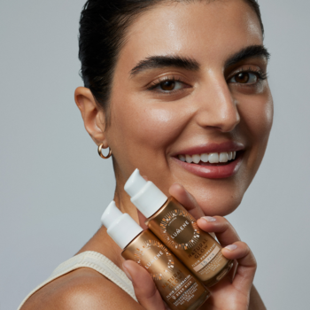 Kobieta uśmiecha się, trzymając dwa podkłady Lumene Natural Glow 1.5 Fair z SPF 20, złote butelki.