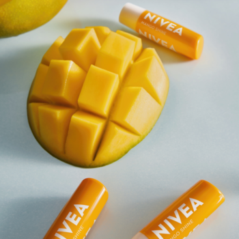 Trzy pomadki NIVEA Mango Shine z masłem shea obok pokrojonego mango na błękitnym tle.