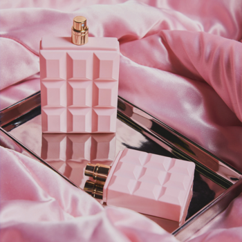 Różowe, teksturowane butelki perfum Sabrina Carpenter Sweet Tooth, większa stojąca, na srebrnej tacy i satynie.