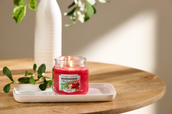 Yankee Candle o zapachu żurawinowym w szklanym słoiczku, z przodu, płonący knot, na białej tacce na drewnianym stole.