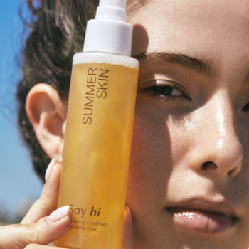 Kobieta trzyma rozświetlającą mgiełkę samoopalającą Say Hi Summer Skin w przezroczystej butelce z bursztynowym płynem.