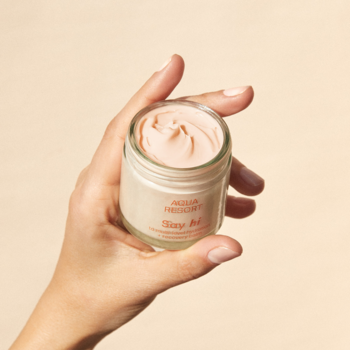 Zbliżenie: krem do twarzy Say Hi Aqua Resort, jasnopomarańczowy, w szklanym słoiczku trzymanym w dłoni.