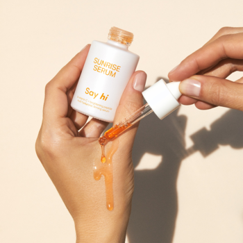 Say Hi Sunrise Serum, biała butelka z pomarańczowym rozjaśniającym serum z witaminą C, dłonie aplikują.