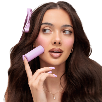 Kobieta aplikuje jasnofioletowy róż w sztyfcie Everybody London Silky Ribbon na policzek. Kremowa formuła, świeży look.