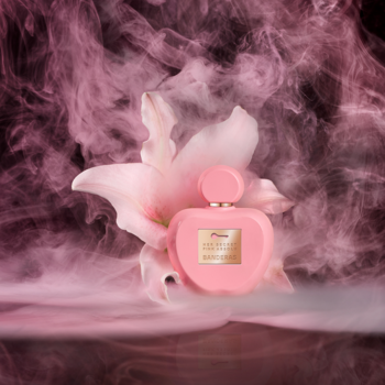 Różowa, jabłkowa butelka perfum Banderas Her Secret EDP otoczona różową mgłą i lilią, widok z przodu.