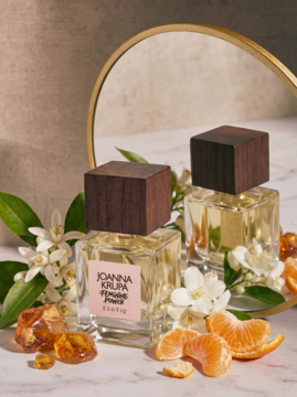 Dwie butelki wody perfumowanej Joanna Krupa Feminine Power Esotiq z drewnianymi korkami, otoczone mandarynkami i kwiatami, przed lustrem.