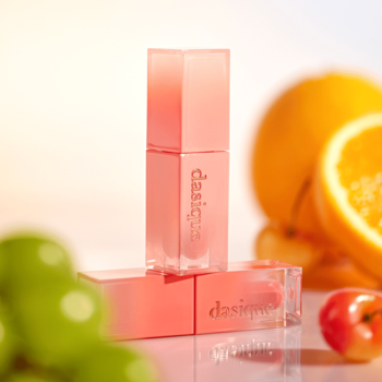 Trzy błyszczyki Dasique Juicy Dewy w koralowych tubkach, ułożone wśród rozmytych owoców cytrusowych i jabłka.
