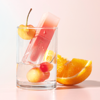 Dasique Juicy Dewy Rose Apple, różowy błyszczyk w szklance z żółtymi wiśniami i pomarańczą, na gradientowym tle.