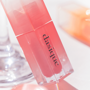 Dasique Juicy Dewy błyszczyk Fig Berry, koralowy, z logo, w przezroczystej butelce pod kątem na jasnym tle.